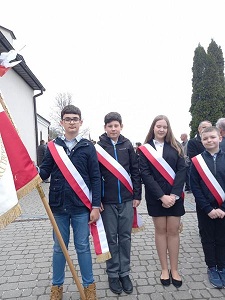 Na zdjęciu znajdują się dzieci z klasy VII i VIII. Trzymają sztandar szkoły, a przez ramię przewieszoną mają biało - czerwoną szarfę